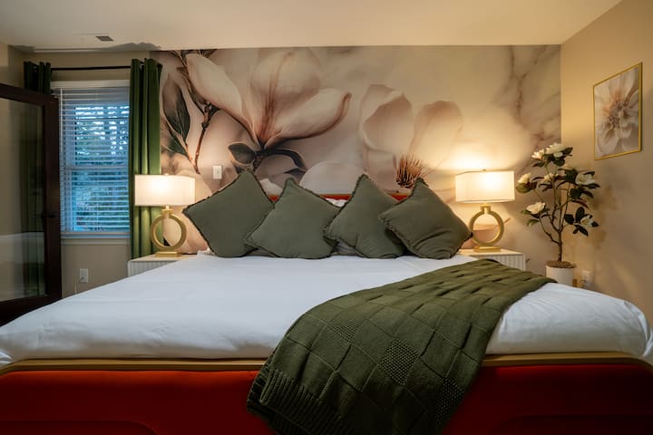 მშვიდ საძინებელში, რომელიც გაწყობილია რბილი საწოლით, მწვანე აქცენტებითა და მაგნოლიის გამოსახულებით დეკორირებული ფოტობუმბულით, იდეალურია დასასვენებლად. ოქროსფერი სანათები და ყვავილებიანი დეტალები ელეგანტურობას სძენს, ბუნებრივი სინათლე და რბილი ტექსტურები კი დამამშვიდებელ გარემოს ქმნის, რომელიც იდეალურია დასვენებისთვის.