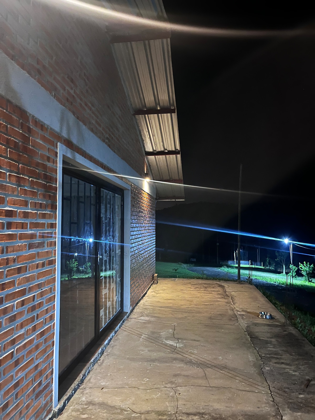 An exterior view of the building is shown at night, with warm lighting illuminating the brick facade. A spacious patio area is visible, featuring a smooth stone pathway, and tall trees can be seen in the background along with distant lighting.