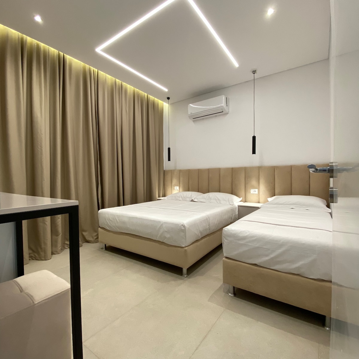 A modern room is illuminated by soft LED lighting. Two spacious beds with neutral-colored upholstery are positioned against a textured wall. Floor-to-ceiling curtains frame a window, while an air conditioning unit is mounted above. A minimalist desk is situated nearby, enhancing the room's functionality.