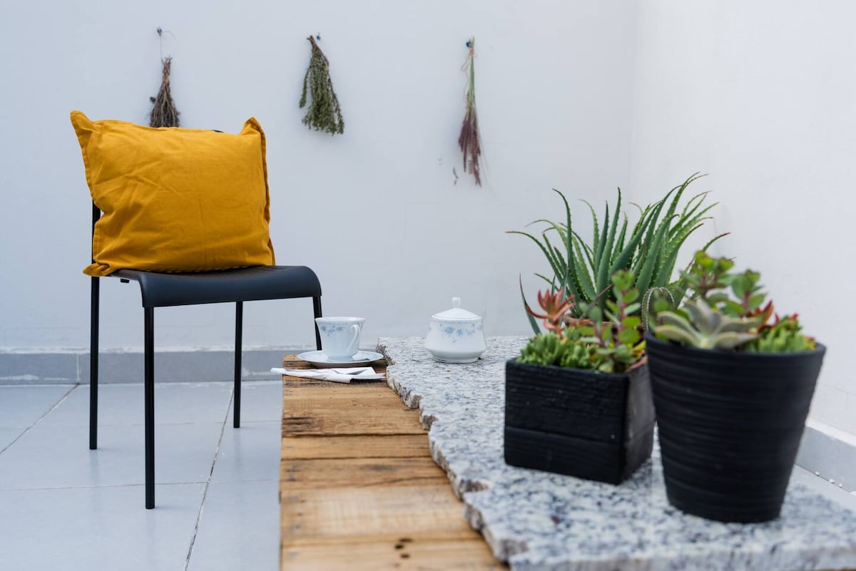 A corner of the outdoor area is presented with a black chair adorned with a yellow cushion. A rustic table made of wood displays two tea cups and a small dish. Succulents in black containers and dried plants hang on the wall, creating a serene environment.