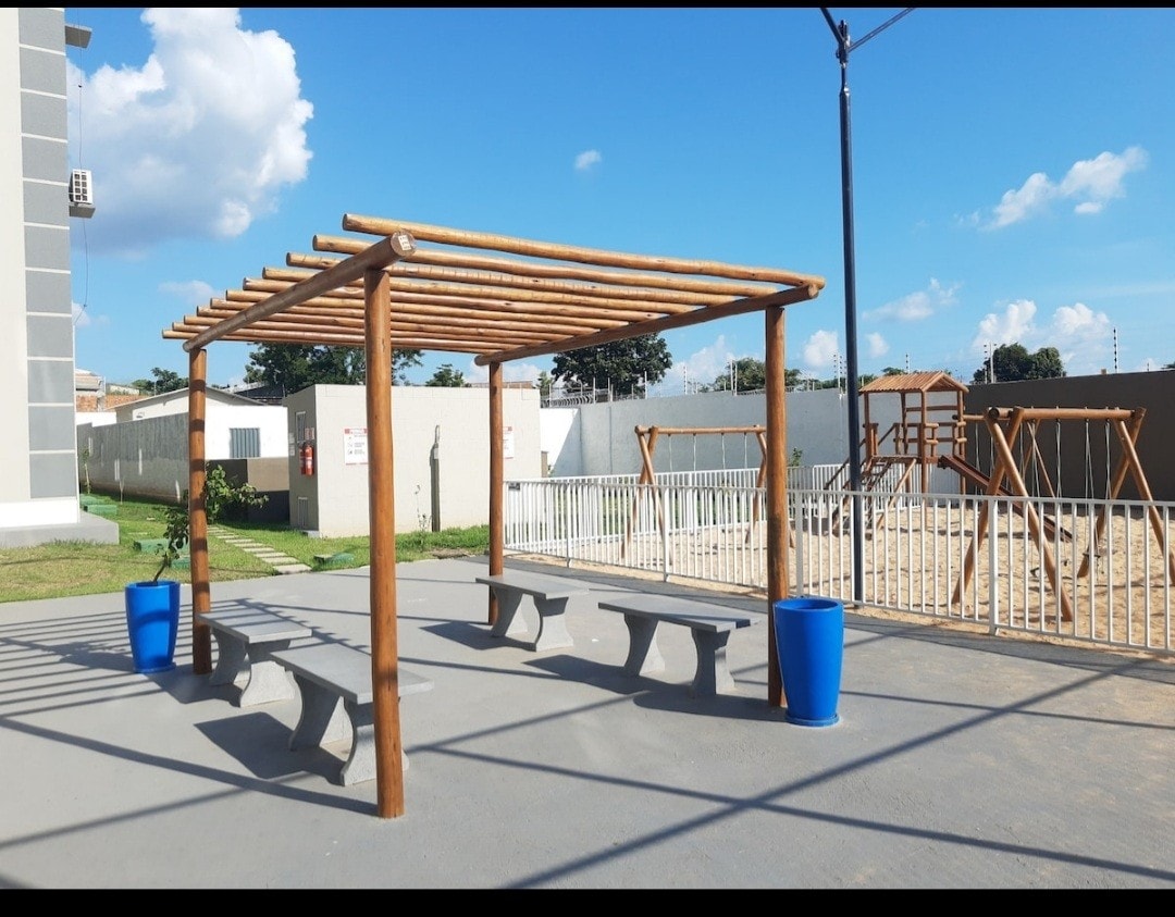 An open outdoor area features a wooden pergola with seating space, surrounded by a smooth, concrete surface. Vibrant blue planters are positioned at either end. In the background, playground equipment can be seen, along with a grassy area and a clear blue sky above.