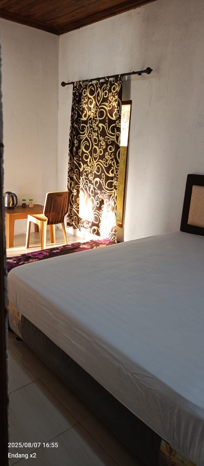 A bright sleeping area features a spacious bed with a light-colored duvet. A window with decorative black curtains allows sunlight to filter softly into the room. A small seating area is visible, including a wooden chair and a kettle on a side table.