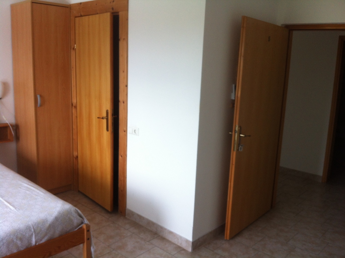A light-filled room is presented with a spacious bed and tasteful wooden furniture. Two closed doors are visible, leading to different areas, while a wardrobe stands beside one door. The tiled floor complements the neutral color palette of the walls.