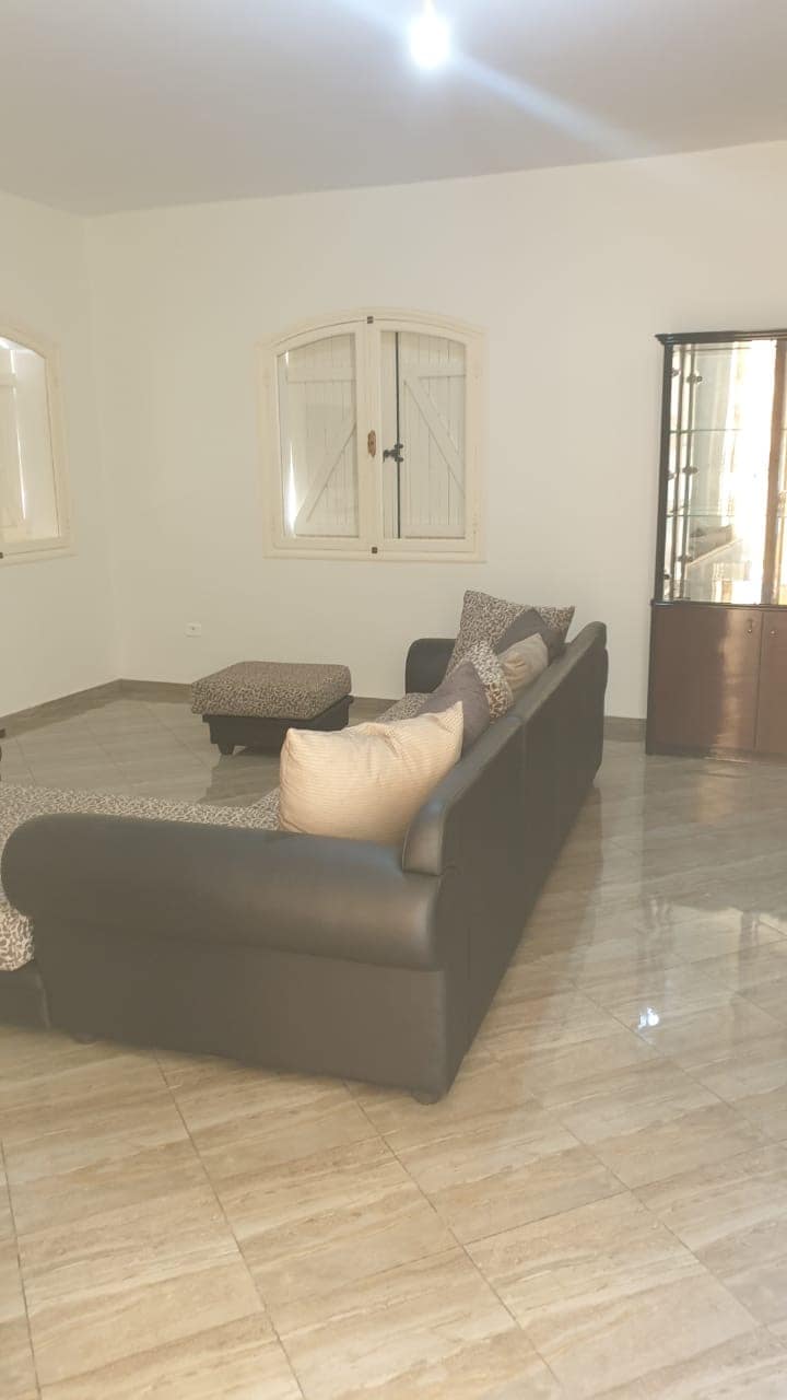 A comfortable seating area is presented, featuring a dark sofa with patterned cushions and an accompanying ottoman. Soft light illuminates the tiled floor, while two windows with shutters allow natural light to enter. A glass cabinet is visible in the corner.