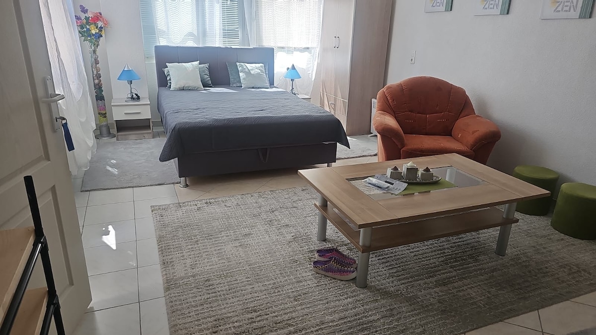 A spacious room features a comfortable double bed with a grey cover and decorative pillows. A cozy orange chair complements a wooden coffee table in the center, while two small green stools are present. The room is well-lit by natural light filtering through sheer curtains.