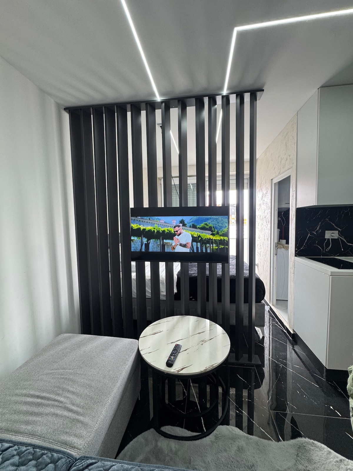 A modern living space is divided by a decorative black slatted partition. A sleek television is mounted on the wall, facing a spacious bed. A light-colored sofa adds comfort, alongside a round table with a remote control on its surface. Natural light illuminates the room.