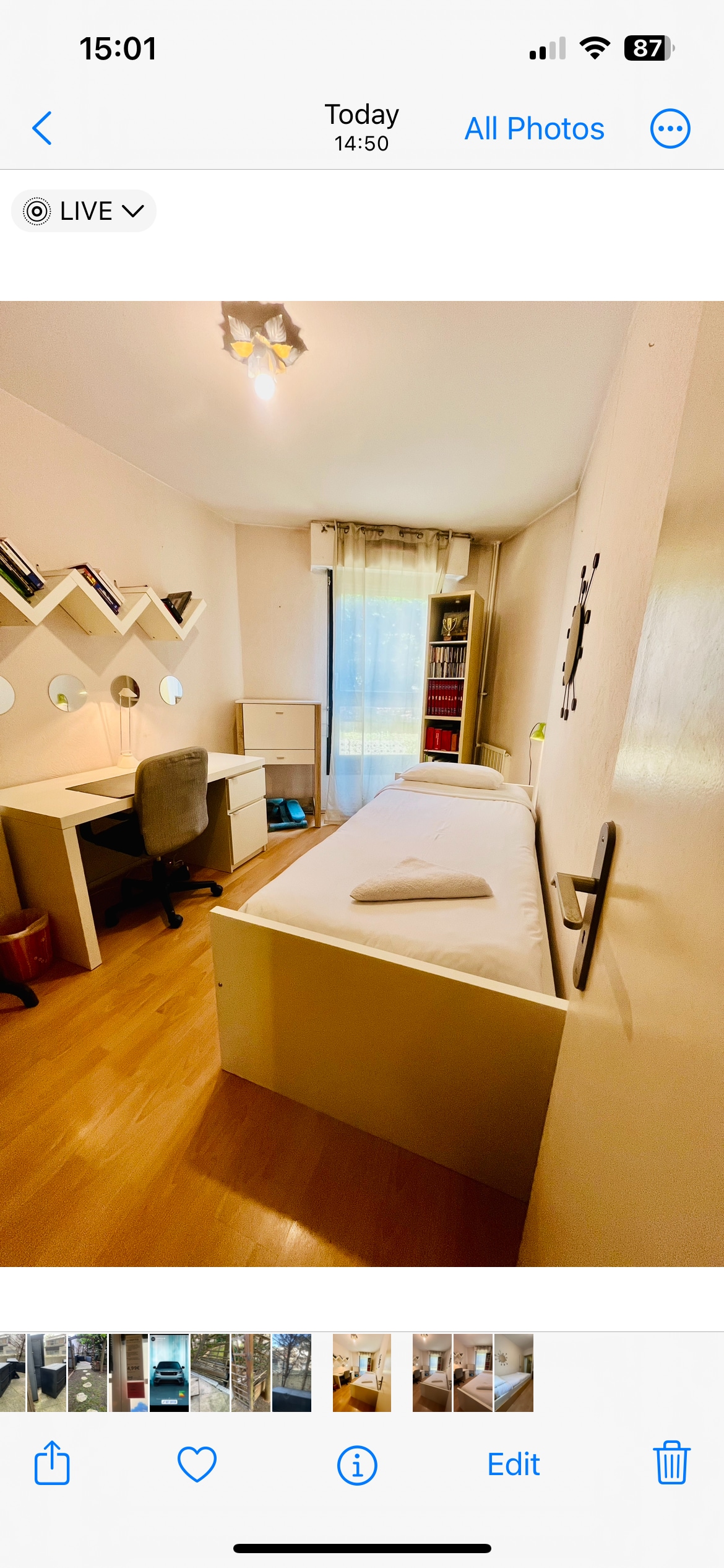 A bright room with natural light entering through a large window, providing a view of gentle curtains. The space features a single bed with a neatly placed towel, a study desk with a comfortable chair, and shelves displaying various items.