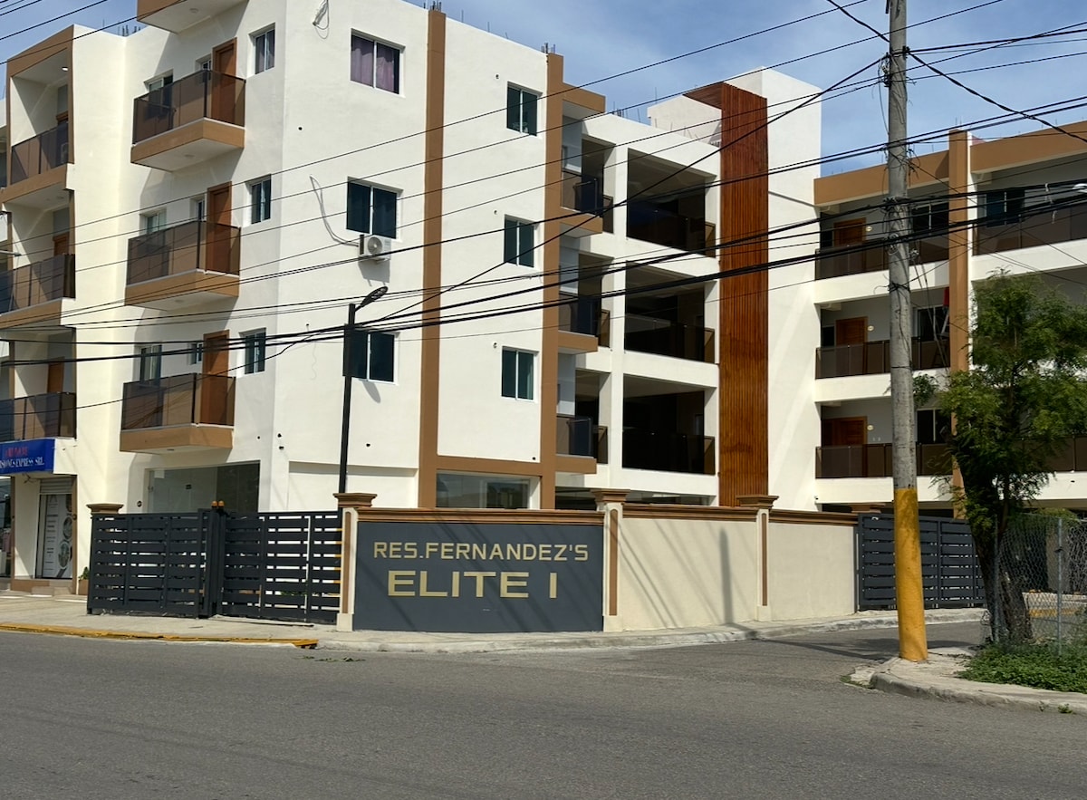 The exterior of a modern apartment building is displayed, featuring multiple balconies and a clean, contemporary design. The entrance is framed by a decorative wall with the name 'ELITE I' prominently featured. Surrounding greenery and urban elements are visible.