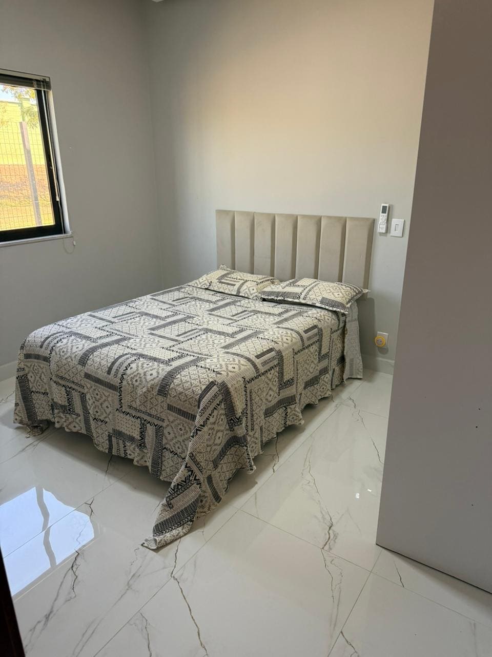 A cozy bedroom features a spacious bed adorned with a patterned blanket. Natural light enters through a window, illuminating the sleek tiled floor. Our welcoming space offers a calming atmosphere, perfect for relaxation after a day of festivities.