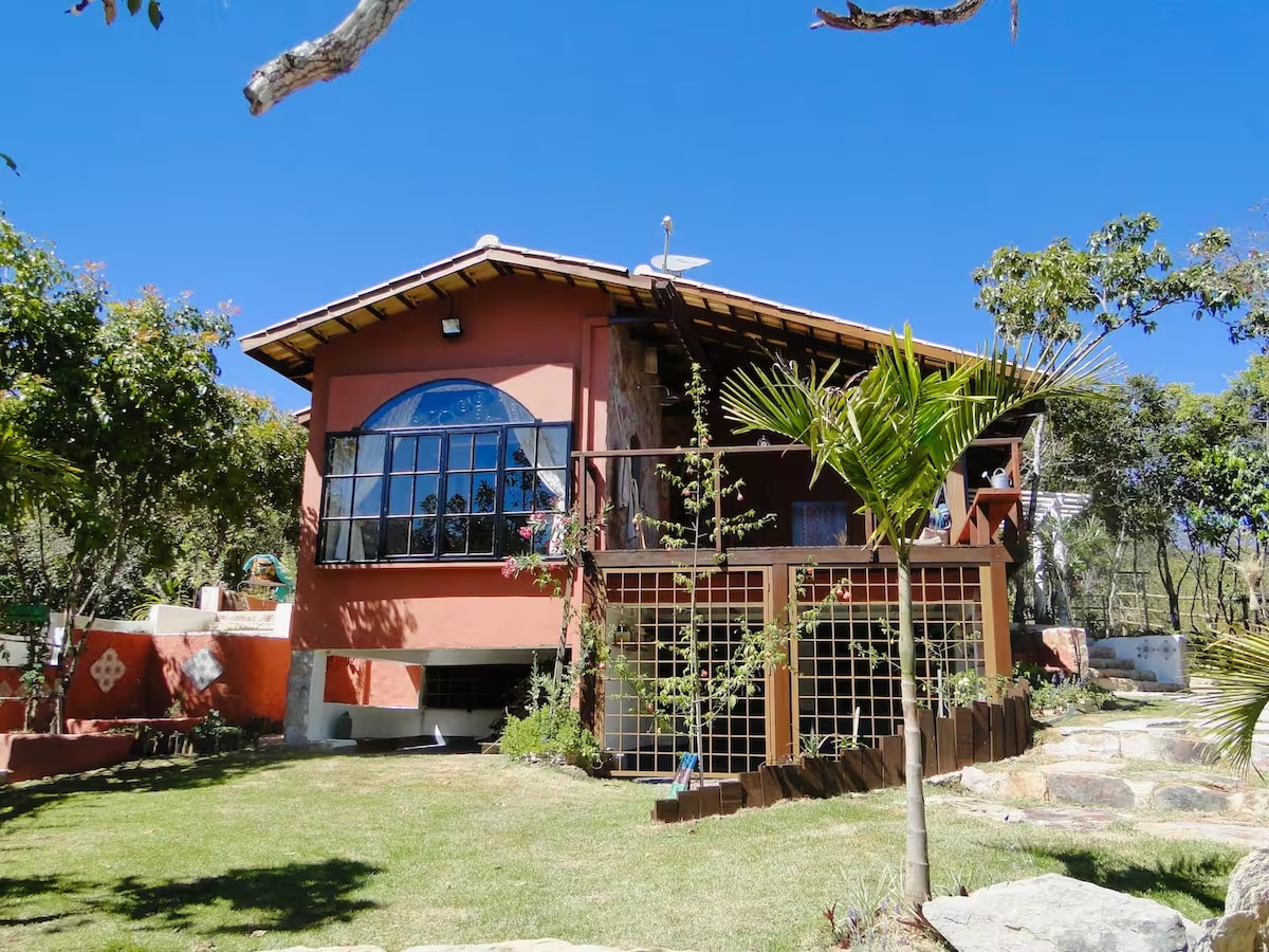 The exterior of a vibrant house is shown, with large windows allowing natural light to enhance the inviting space. Lush greenery surrounds the property, complemented by a well-maintained lawn. A deck area provides outdoor living space, offering a glimpse of the tranquil environment.
