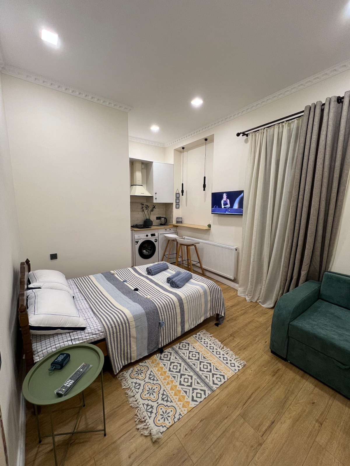 A compact living area features a bed with a striped blanket, a small green side table, and a couch in muted tones. A kitchenette is visible with a washing machine, and large curtains frame the window, allowing light to enhance the space.