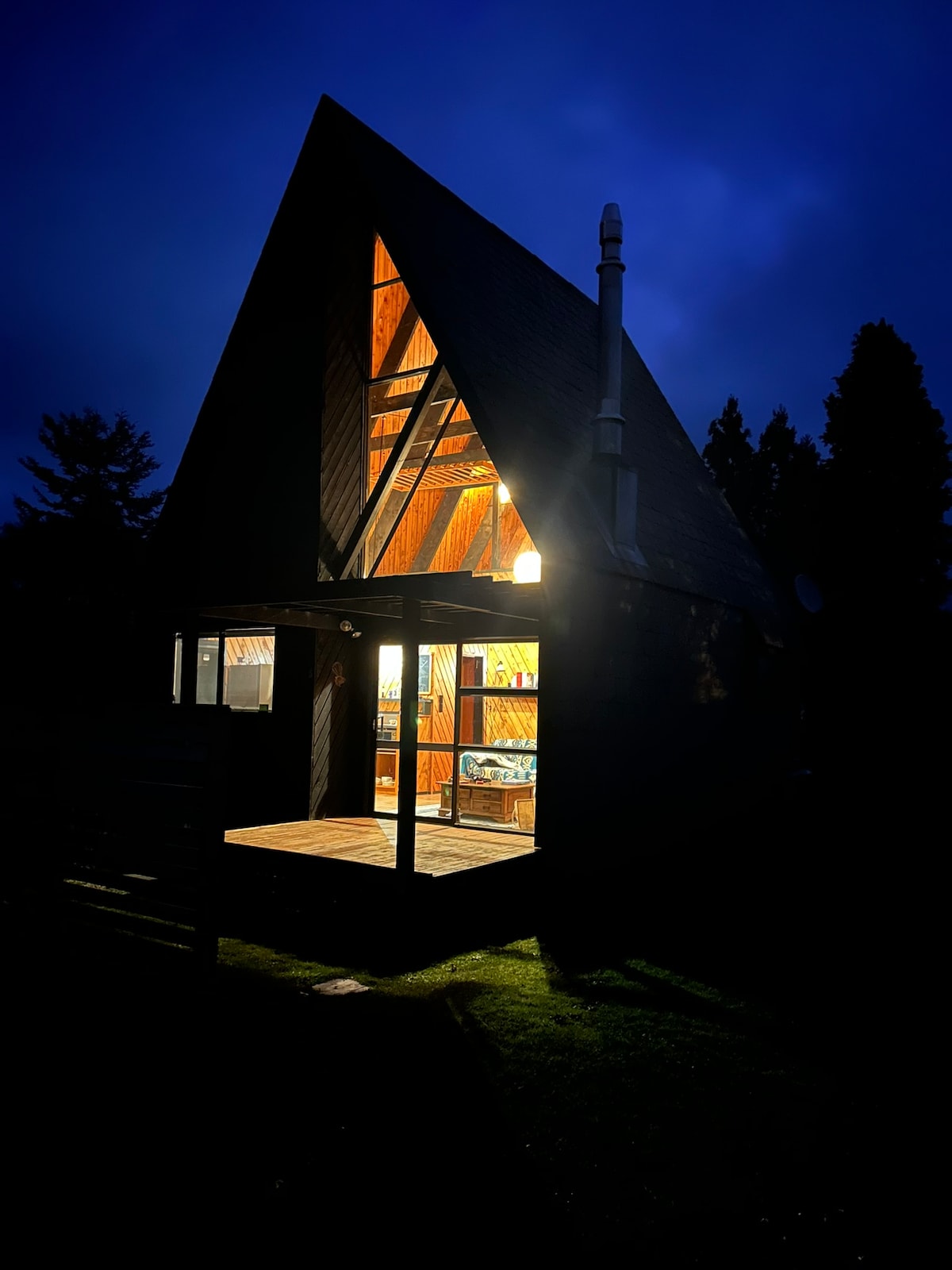 An A-frame chalet is illuminated at night, showcasing warm light spilling from its open entrance. The structure features a steeply pitched roof and large windows, emphasizing its architectural charm against a dark, cloudy sky. Nearby greenery adds a touch of nature.