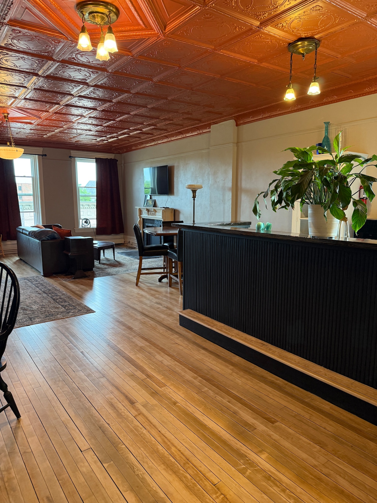 The living area is highlighted by rich wooden flooring and a striking orange tin ceiling. A comfortable seating area features a dark sofa and a wooden coffee table, while a small desk and chair are positioned nearby. Natural light floods the space through large windows draped with dark curtains.