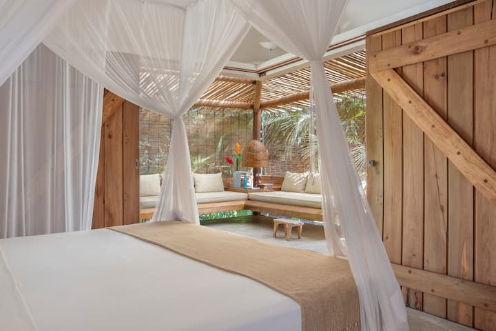 Въздушна стая с легло, увито в светла завеса, дървени врати в рустик стил и директна връзка с открития салон – убежище, което съчетава комфорт и природа.