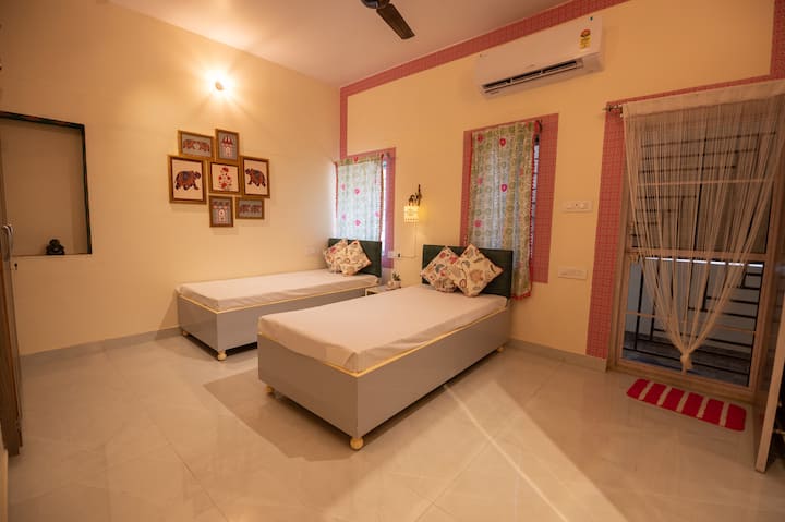 La chambre à coucher dégage un charme chaleureux du Rajasthan avec des lits simples jumeaux, des coussins imprimés en bloc vibrants et des œuvres d'art murales traditionnelles. Profitez du confort de la climatisation, de l'éclairage apaisant, de la salle de bain privée et de la garde-robe, parfaits pour les voyageurs à la recherche de style et d'élégance.