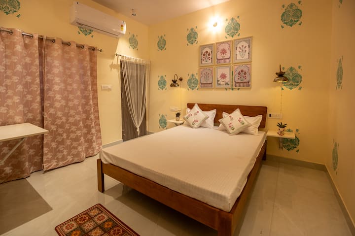 Cette chambre dégage une élégance calme du Rajasthan avec un lit queen confortable, des murs à imprimé floral et des rideaux pastel doux. Les œuvres d'art murales artisanales et les lampes suspendues dorées ajoutent un charme royal. Détendez-vous dans le confort climatisé et l'ambiance sereine et apaisante.