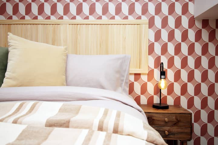 Un dormitorio boutique bellamente diseñado con una lujosa cama doble colocada contra una llamativa pared geométrica. La suave iluminación ambiental, la lujosa ropa de cama y los cálidos detalles de madera crean un refugio tranquilo y relajante.