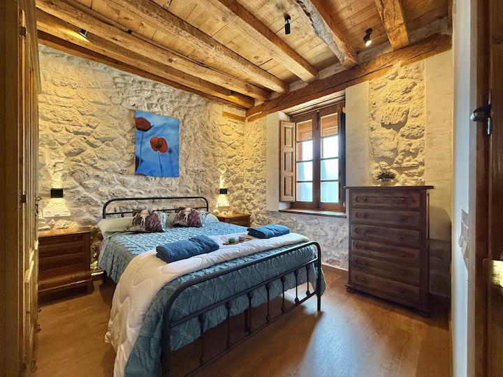 חדר שינה עם מיטה זוגית, קירות אבן ותקרת עץ שיוצרים אווירה חמה ואותנטית. מושלם למנוחה וליהנות מהשלווה הכפרית.