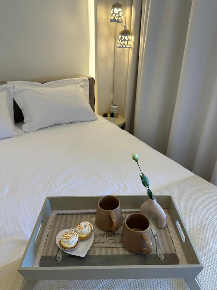 חדר שינה מודרני ומינימליסטי עם תאורה רכה. מיטת קינג סייז מוצעת במרכז החדר עם מצעים לבנים, מול קיר מואר. מנורות תליון ושידות לילה קטנות מוסיפות מגע נעים. ארון בגדים עם 4 דלתות מוסיף מגע נעים.  