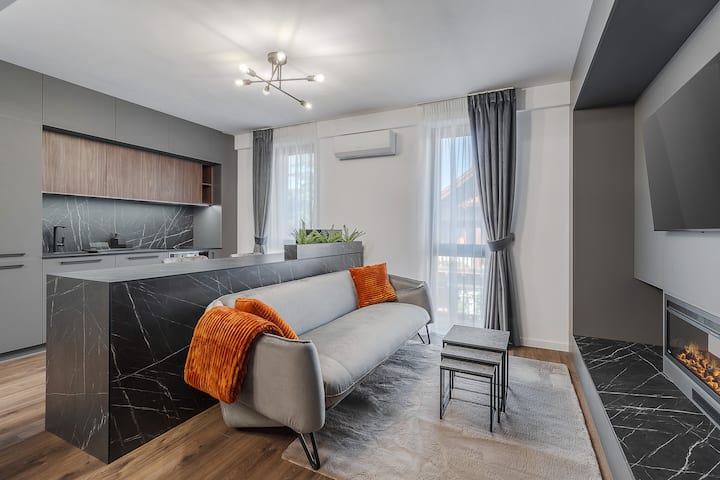 La refinada sala de estar cuenta con una moderna chimenea eléctrica, TV inteligente y un sofá extremadamente cómodo, creando un ambiente premium. La cocina gourmet totalmente equipada completa este amplio espacio, ideal para una relajación absoluta en un entorno lujoso.