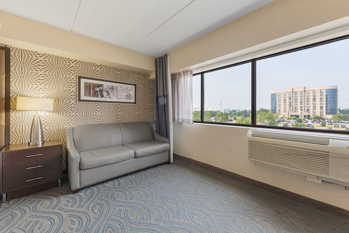 Მყუდრო მოსასვენებელი სივრცე გლუვი ტყავის თეთრეულით და დიდი ფანჯრით, სადაც ბევრი ბუნებრივი სინათლეა. Დეკორში არის თანამედროვე ლამპა და კედლის ელეგანტური ხელოვნება.