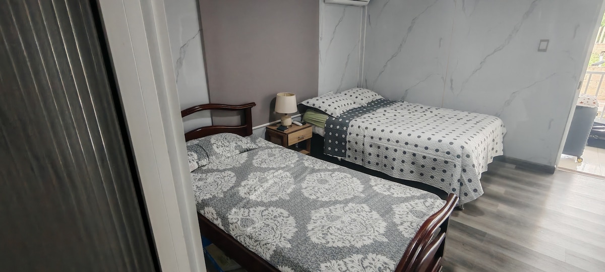A tranquil sleeping area is presented with two beds, one double and one single, covered with patterned bedding. A bedside table with a lamp is located beside the double bed. Light-colored walls and wooden flooring enhance the clean and comfortable ambiance.