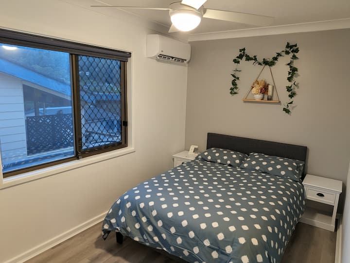 เตียงคู่พร้อมโต๊ะข้างเตียงและโคมไฟ นอกจากนี้ยังมีแท่นชาร์จไร้สาย พัดลม เครื่องปรับอากาศ และตู้เสื้อผ้าพร้อมกระจกและไม้แขวนเสื้อ 