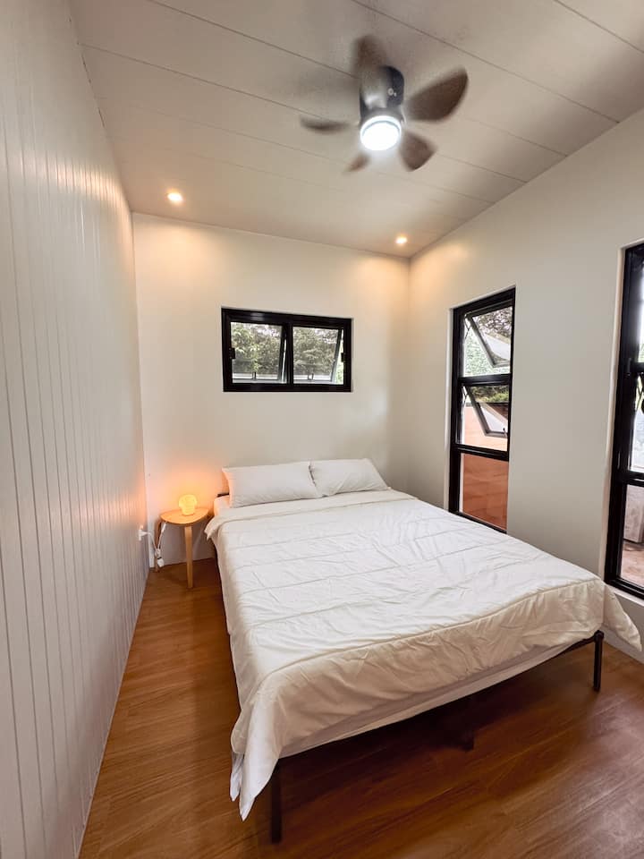 El dormitorio cuenta con una cómoda cama tamaño queen con sábanas limpias e iluminación suave. Las grandes ventanas dejan entrar luz natural y brisa fresca, mientras que el ventilador de techo mantiene el espacio aireado y relajante.