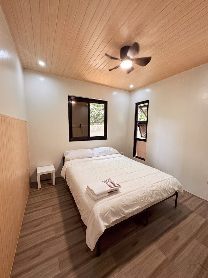 El dormitorio cuenta con una cómoda cama tamaño queen con sábanas limpias e iluminación suave. Las grandes ventanas dejan entrar luz natural y brisa fresca, mientras que el ventilador de techo mantiene el espacio aireado y relajante.
