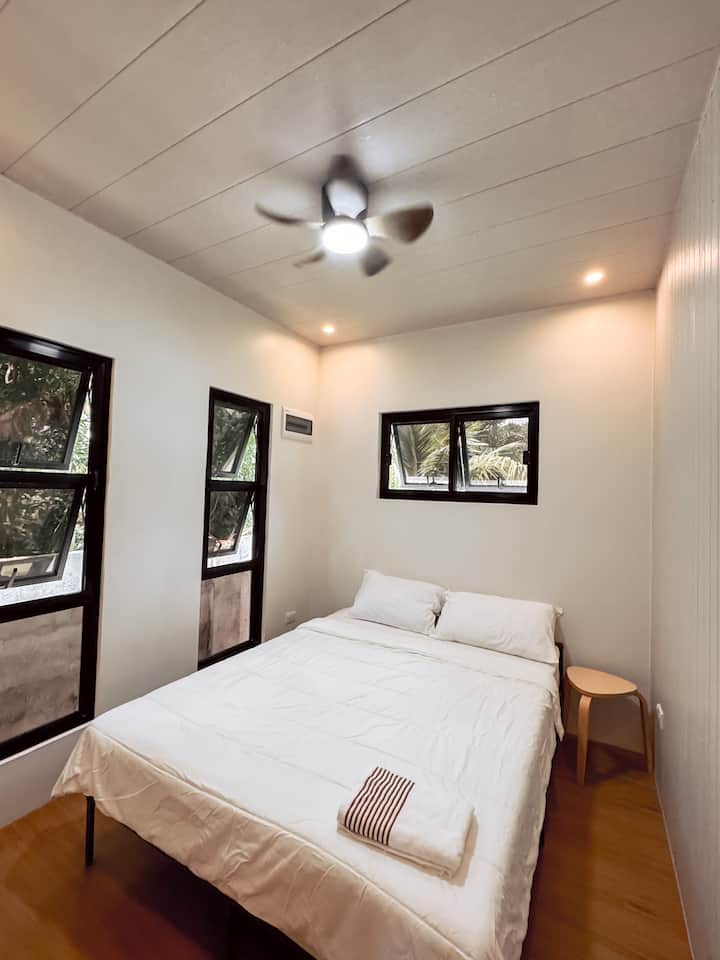 El dormitorio cuenta con una cómoda cama tamaño queen con sábanas limpias e iluminación suave. Las grandes ventanas dejan entrar luz natural y brisa fresca, mientras que el ventilador de techo mantiene el espacio aireado y relajante.