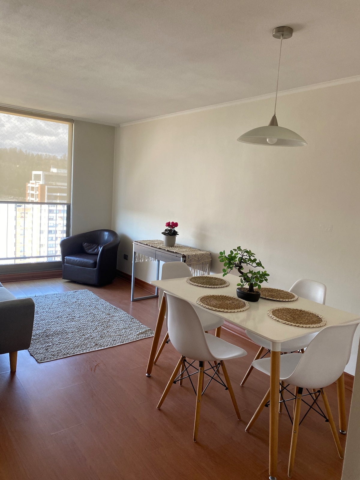 A well-lit dining area features a white table surrounded by four chairs. A small potted plant is placed at the center. A cozy armchair and a textured area rug complement the space, with a large window providing sunlight and showcasing distant mountain views.