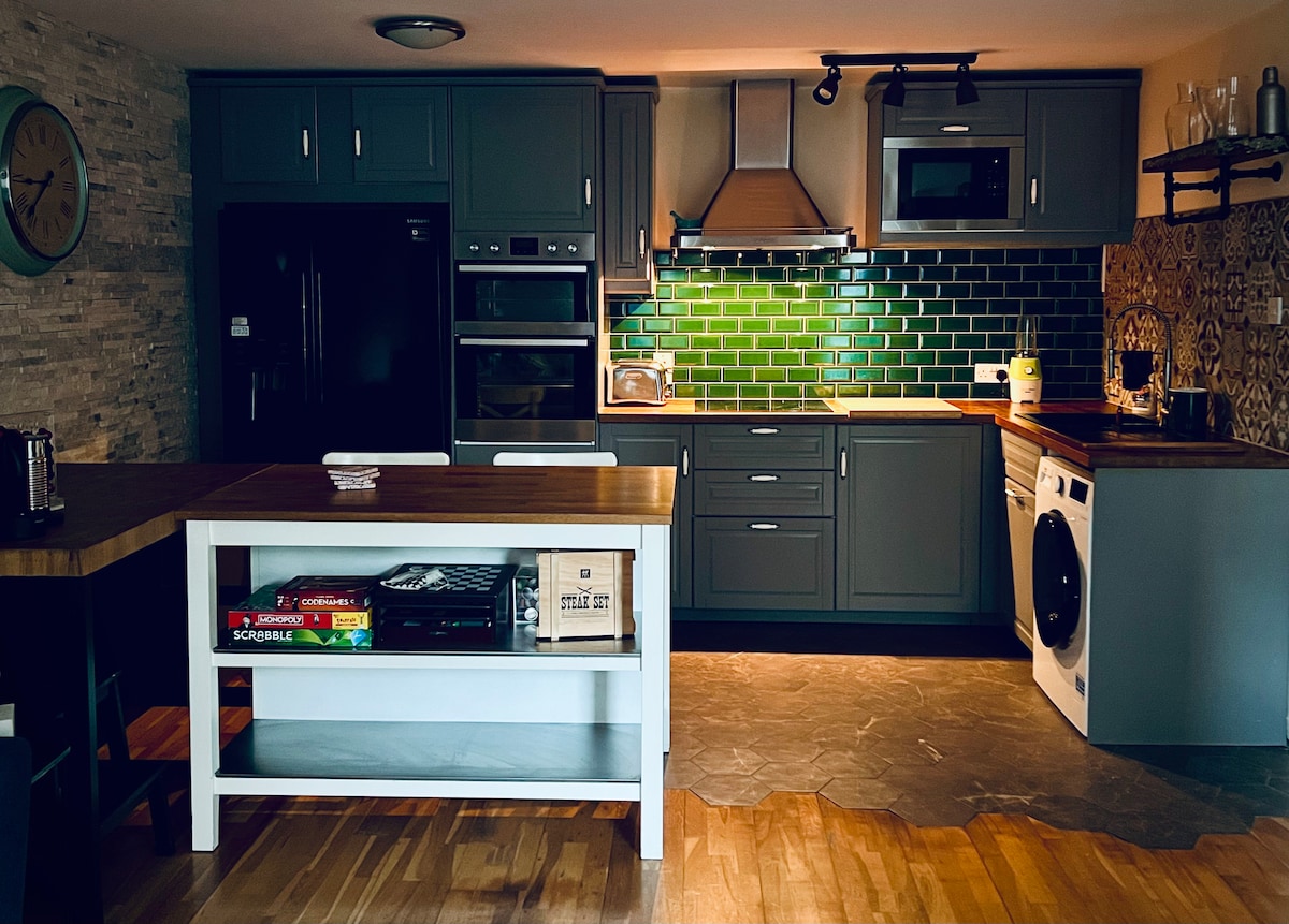 A well-equipped kitchen features grey cabinetry with a contrasting green tile backsplash. Modern appliances, including a stove and refrigerator, are visible alongside an island with a wooden countertop. A mix of textures, such as stone flooring and decorative wall elements, adds depth to the space.