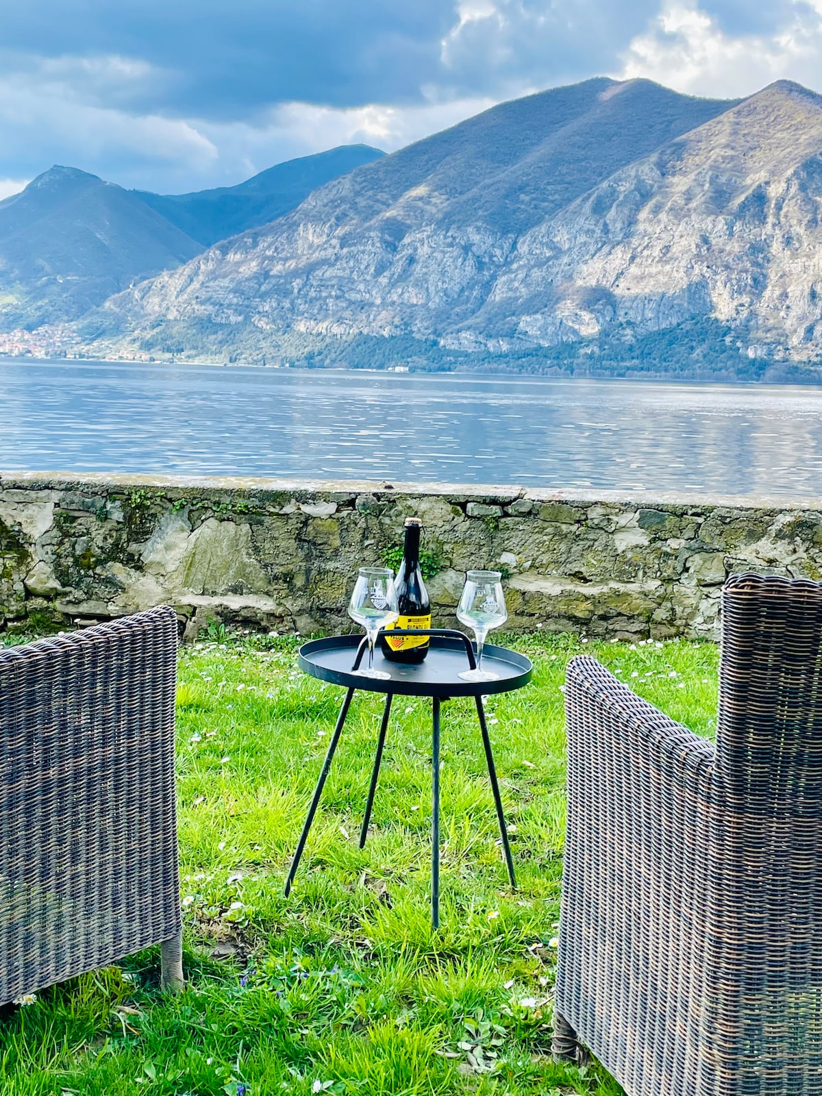 A serene outdoor seating area is set against a backdrop of the lake and mountains. Two wicker chairs surround a small black table topped with a bottle of champagne and three glasses. The lush green grass enhances the scenic view of the water and surrounding landscape.
