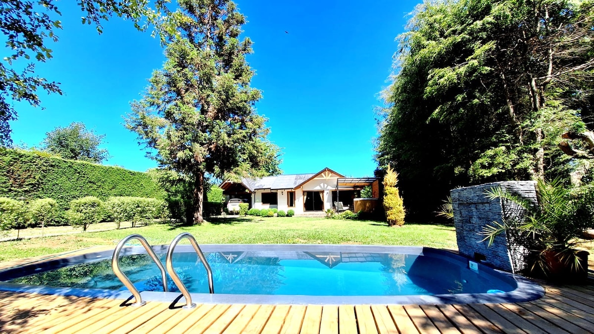 A private swimming pool is set within a wooden deck, surrounded by lush greenery and tall trees. The cabin, featuring a spacious front yard, is visible in the background under a clear blue sky.
