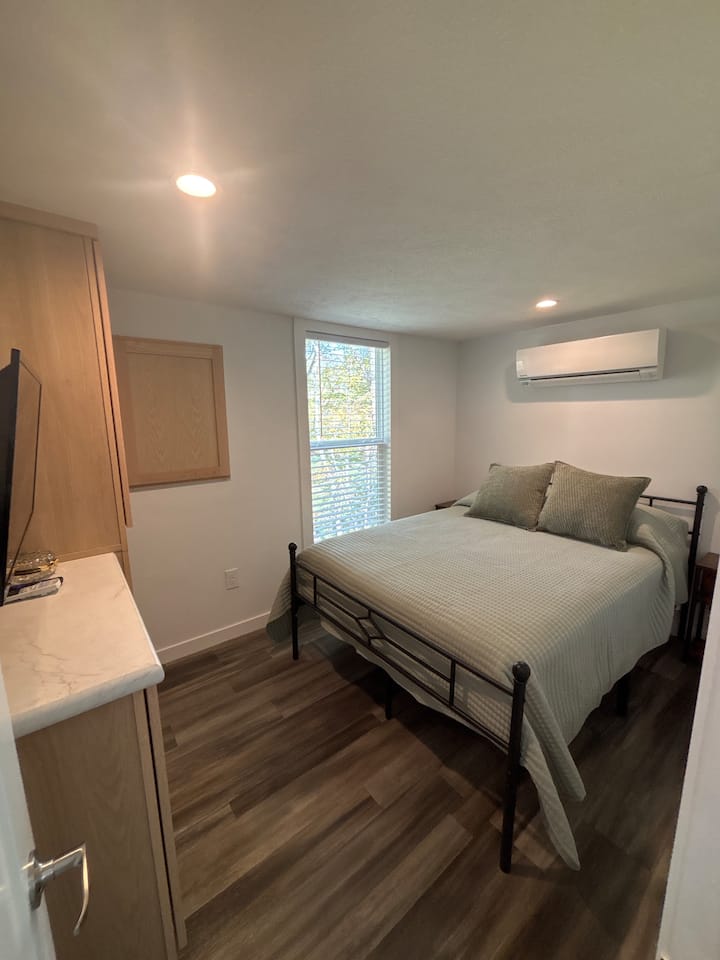 Cozy bedroom with a full-size bed, soft linens, and extra pillows. Features a mounted TV, built-in storage, and large windows that let in plenty of natural light — a perfect spot to relax after a day exploring the mountains or wandering the farm.