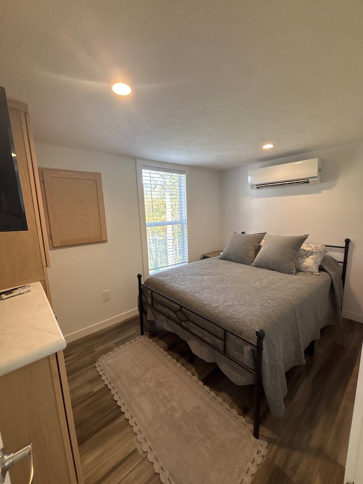 The bedroom at Carolina Chickadee Cottage features a cozy queen bed with soft linens, a mounted TV, built-in storage, and large windows that let in plenty of natural light—creating a calm, comfortable space to rest after a day in the mountains.