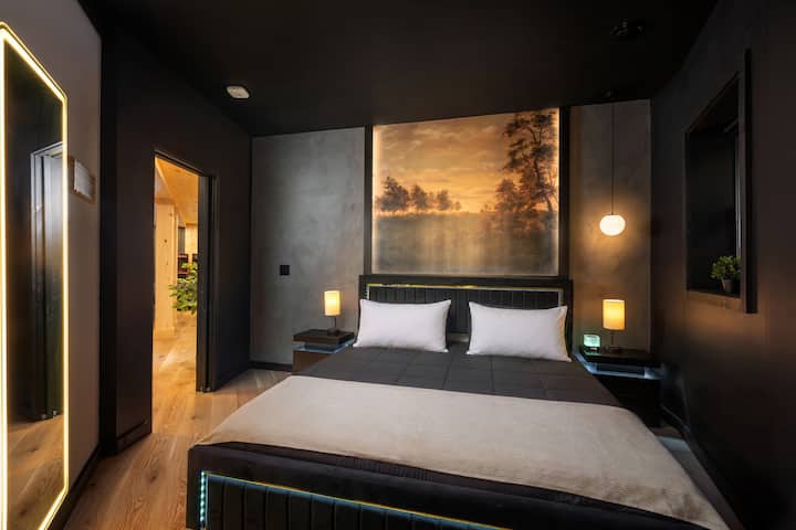 Bedroom 4 with a king-size bed, dark modern walls, warm bedside lighting, large scenic wall art, illuminated mirror, and a cozy luxury cabin-style design.