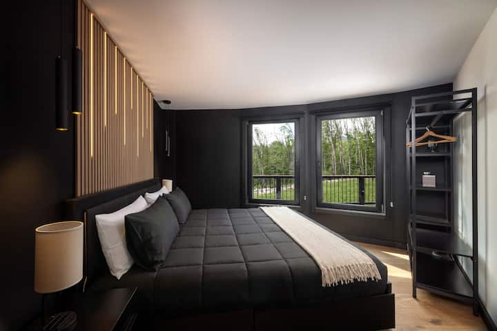 Bedroom 3 with a king-size bed, modern black accent wall, wood slat detail, bedside lighting, open closet shelving, and large windows with wooded views.
