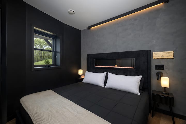 Bedroom 6 with a king-size bed, dark modern walls, gray accent wall, warm LED lighting, bedside lamps, and a window overlooking the peaceful wooded scenery.