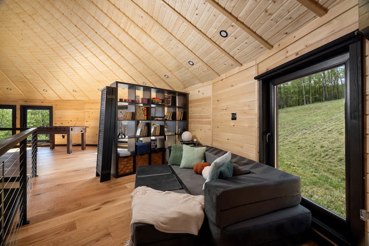 Loft sofa area with a cozy sectional, large picture window, built-in bookshelf, open railing, and peaceful wooded views, creating a relaxing upstairs lounge space.