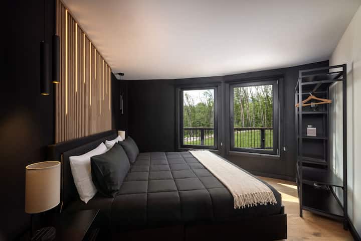 This bedroom pairs a cozy bed with corner windows that open onto the forest and deck, plus an open clothing rack for easy unpacking on longer stays.