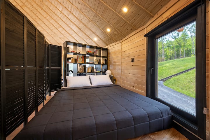 Extra loft bedroom with wood-paneled walls, a king sofa bed, and a large window overlooking the wooded outdoor scenery.