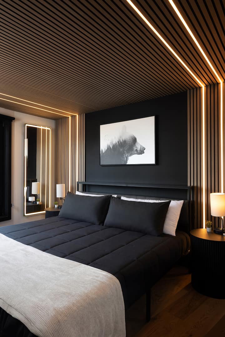 Bedroom 1 with a king-size bed, modern black walls, warm wood slat ceiling accents, soft LED lighting, bedside lamps, and a cozy luxury cabin feel.