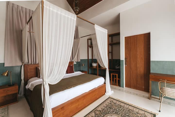 Amplio dormitorio de Bali con cama de madera con dosel, cortinas blancas fluidas y decoración terrosa. Las paredes con detalles en verde suave, las estanterías integradas y un escritorio crean un ambiente sereno de villa boutique perfecto para relajarse.