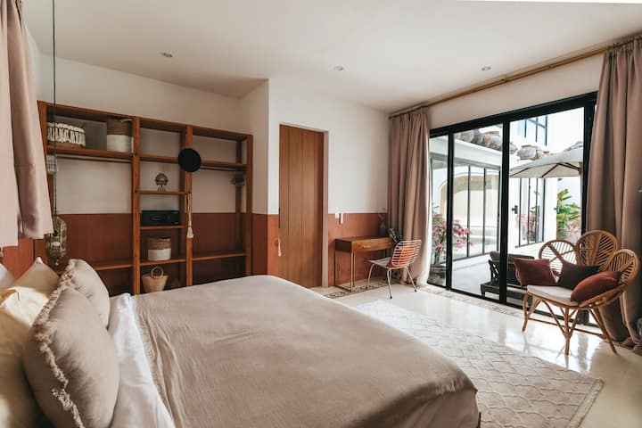 Dormitorio cálido y espacioso con una cama tamaño king, estanterías abiertas de madera y un acogedor espacio de trabajo. Las cortinas del piso al techo y las puertas corredizas se abren a la piscina, creando un refugio de villa de Bali brillante y sereno perfecto para relajarse y descansar.