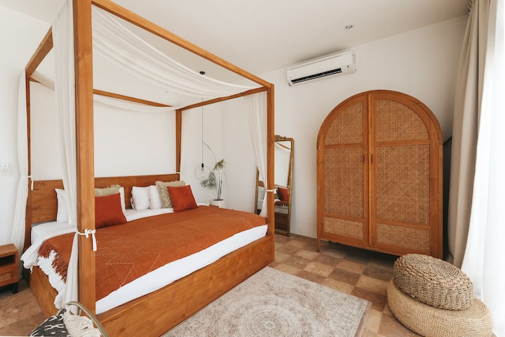Dormitorio luminoso de inspiración balinesa con cama con dosel de madera, ropa de cama de terracota en tonos tierra y un gran armario de ratán.Las suaves cortinas drapeadas, las texturas naturales y un espejo vintage crean un refugio sereno de estilo boutique.