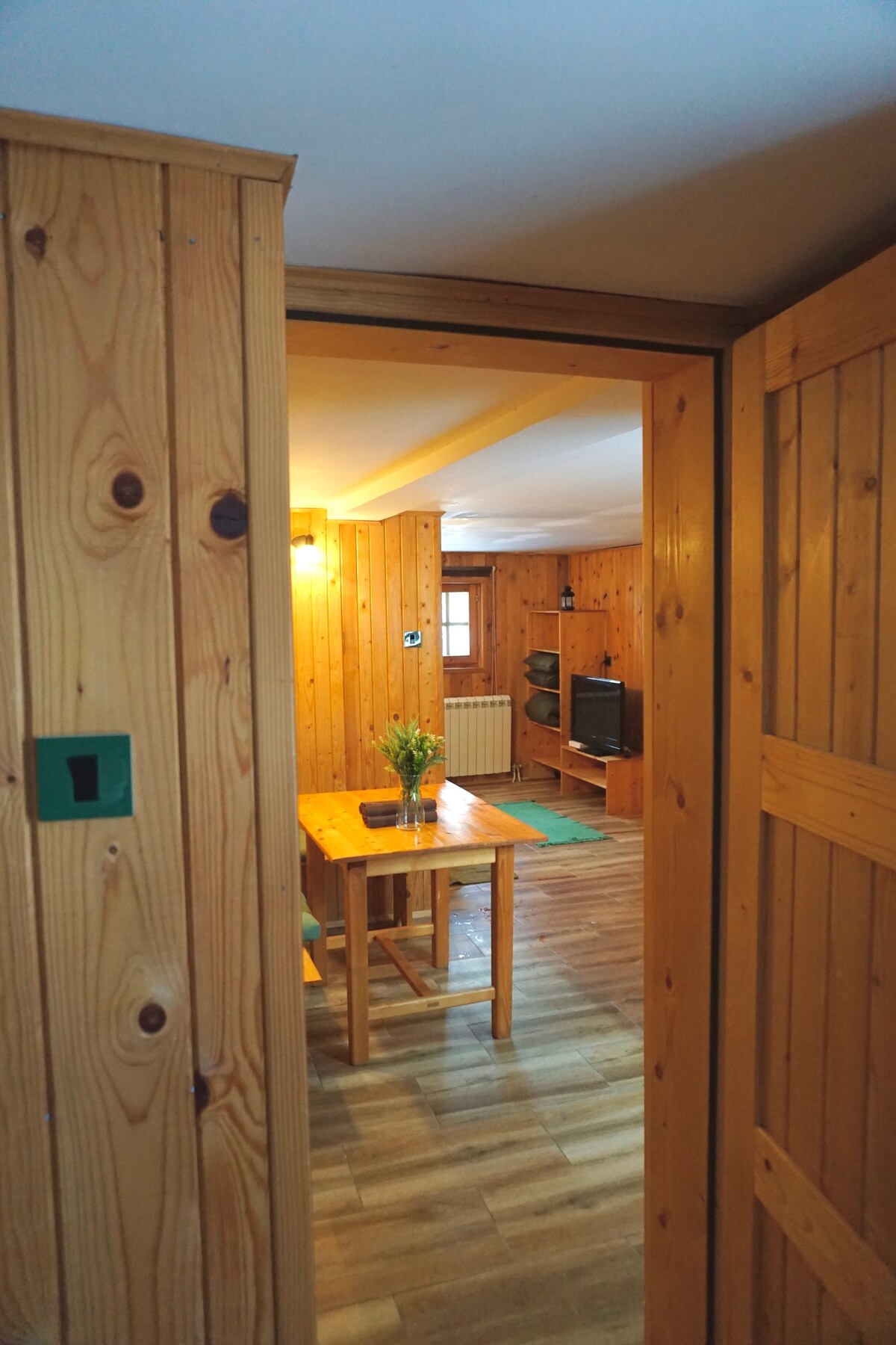 A wooden interior is visible upon entering, featuring a cozy living room area with a dinette table and chairs. Natural wood paneling adorns the walls, and a television is positioned against one wall. Soft lighting enhances the inviting environment.