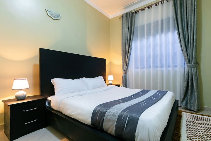 Lit confortable grand lit ou très grand lit avec des draps blancs frais, un couvre-lit gris et une tête de lit noire. Une grande fenêtre avec des rideaux laisse entrer la lumière naturelle, créant une ambiance lumineuse et aérée.