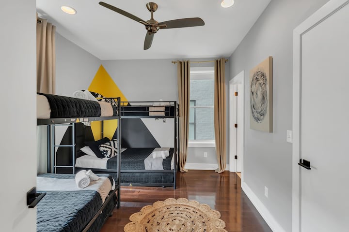 Bright bunk room with two modern twin-over-twin beds, graphic accent wall, and coordinated bedding. Large window and ceiling fan keep the space airy, making it ideal for kids or group stays. Two additional twin rollout beds.