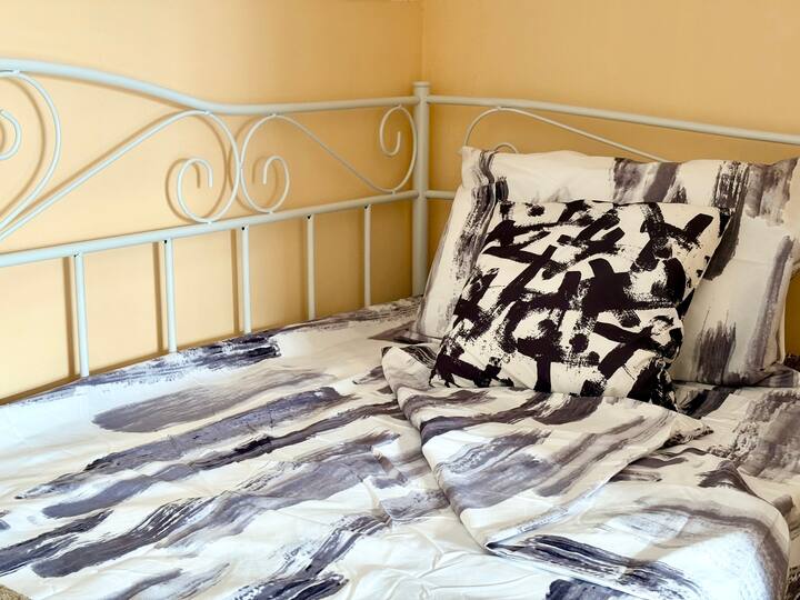 غرفة نوم في الطابق الثاني مع سريرين مفردين وتلفزيون ومكيف هواء. تملأ النوافذ الكبيرة الغرفة بالضوء الطبيعي، مما يخلق مساحة مشرقة ومريحة للراحة والاسترخاء.